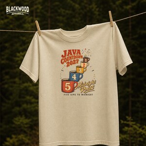 Può includere: Una t-shirt color beige con una grafica di cinque tazze da caffè impilate numerate da uno a cinque. Il testo recita "JAVA COUNTDOWN 2027", "Midnight Roast" e "FIVE SIPS TO MIDNIGHT". Il logo "BLACKWOOD APPARELS." è stampato in alto a sinistra.