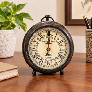 Puede incluir: Un reloj de mesa redondo de metal en tono bronce oscuro con esfera color crema, números romanos negros y un icono de barco. El texto Jefferson & Smith London está impreso en la esfera. Tiene un asa superior y dos pies pequeños.