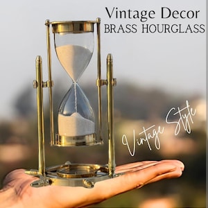 May include: A brass hourglass with clear glass bulbs containing white sand, held within a rotating metal frame. The text "Vintage Decor BRASS HOURGLASS" and "Vintage Style" is visible in the upper right and lower right corners of the image.