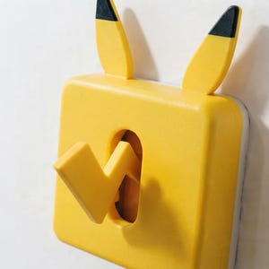 Puede incluir: Una cubierta de interruptor de luz amarilla con dos orejas puntiagudas con puntas negras en la parte superior y un interruptor de palanca en forma de rayo en el centro. La placa cuadrada es de color amarillo brillante.