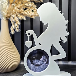 May include: A white silhouette figure of a pregnant person standing on an oval base. The figure features a circular cutout at the abdomen area containing a black and white ultrasound image and a small heart-shaped charm hanging from one hand.