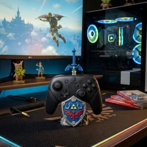May include: A black wireless game controller resting on a stand shaped like a blue and silver shield with a red bird emblem. A blue sword replica is inserted into the top of the controller. The stand features a gold triangular triforce symbol.