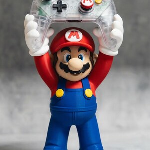 May include: A Mario figurine wearing a red hat with an M emblem, a red shirt, blue overalls with yellow buttons, and brown shoes. The figure holds a clear, transparent video game controller above its head with both white-gloved hands.