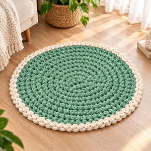 Puede incluir: Una alfombra redonda tejida a mano que presenta un centro texturizado de color verde salvia hecho con hilo grueso. La alfombra está terminada con un borde ancho y trenzado en color blanco crema, creando un contraste entre las secciones.