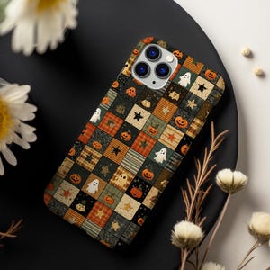 Può includere: Una custodia per smartphone con un motivo patchwork a quadrati nei toni dell'arancione, nero e beige. Il design presenta motivi ripetuti di zucche, fantasmi bianchi e stelle nere, con un'apertura per il sistema a tre fotocamere.