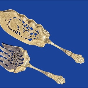 May include: A set of two silver-plated serving pieces with intricate cut-out designs. The pieces are a serving fork and a serving spoon, both with ornate handles.