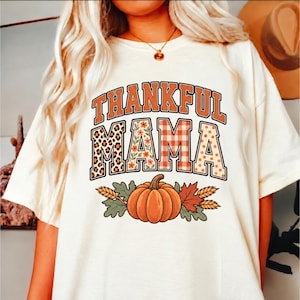 Op de afbeelding: Een crèmekleurig T-shirt met korte mouwen en de tekst THANKFUL MAMA in oranje blokletters. De letters bevatten luipaardprint, bloemen, ruitjes en stippen. Onder de tekst staat een oranje pompoen met groene bladeren.