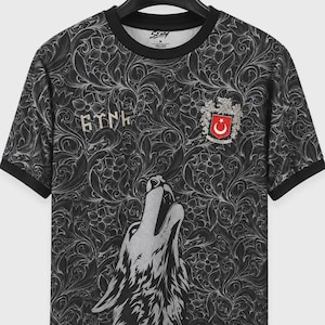 Może przedstawiać: Czarny t-shirt z krótkim rękawem, ozdobiony skomplikowanym szarym wzorem kwiatowym, grafiką wyjącego wilka u dołu oraz napisem "TÜRK" stylizowanym na runy. Herb z flagą Turcji znajduje się w górnej prawej części klatki piersiowej.