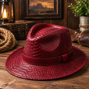 Pode incluir: Um chapéu fedora vermelho escuro com um padrão texturizado de pele de cobra em toda a superfície, incluindo a copa e a aba larga. O chapéu inclui uma faixa de pele de cobra vermelha a condizer que envolve a base da copa do chapéu.