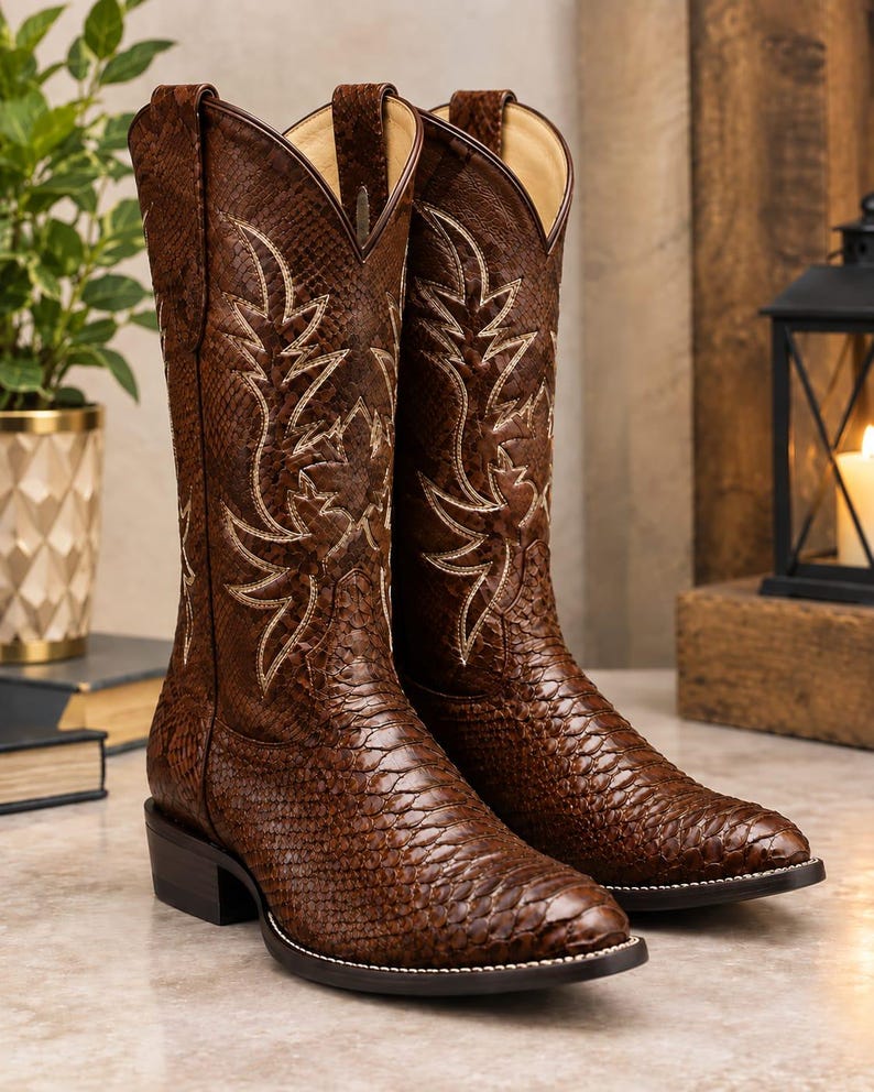 May include: A pair of brown cowboy boots featuring a textured snakeskin-like pattern across the exterior. The boots have a pointed toe, a low block heel, and intricate cream-coloured decorative stitching on the shaft in a leaf-like design.