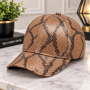 Puede incluir: Una gorra de béisbol con un patrón texturizado inspirado en la piel de serpiente en tonos bronceado y marrón. La gorra tiene una corona estructurada y una visera curva, con un pequeño botón en la parte superior central. El material muestra un diseño.