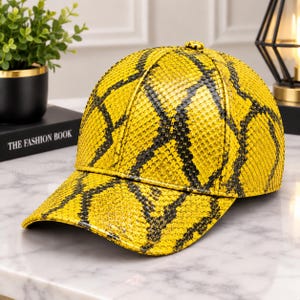 Può includere: Un cappellino da baseball con un vivace motivo a pelle di serpente giallo e nero. Il cappellino presenta una corona strutturata, una visiera curva e un piccolo bottone al centro. Il materiale testurizzato imita l'aspetto delle squame.