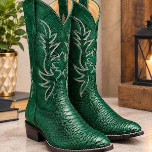 May include: A pair of emerald green cowboy boots featuring a textured snakeskin-like pattern on the foot and shaft. The boots have a pointed toe, a low block heel, and white decorative stitching in a leaf-like pattern on the upper shaft.