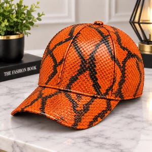 Pode incluir: Um boné de beisebol com um padrão de pele de cobra em laranja vibrante e preto. O boné tem uma coroa estruturada de seis painéis, uma aba curva e um pequeno botão a condizer no topo. A superfície texturizada imita escamas de réptil.
