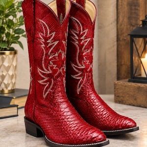 May include: A pair of red cowboy boots featuring a textured snakeskin-like pattern, a pointed toe, and a stacked heel. The shaft of each boot is decorated with intricate white stitching in a leaf-like design and includes two pull tabs at the top.
