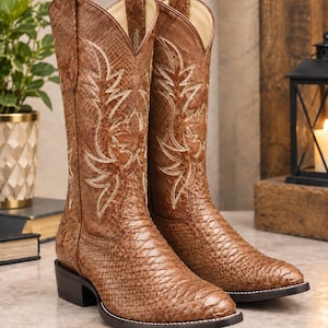 Könnte beinhalten: Ein Paar braune Cowboystiefel mit einem strukturierten Schlangenledermuster auf dem gesamten Leder. Die Stiefel haben eine spitz zulaufende Kappe, einen niedrigen Blockabsatz und dekorative weiße Nähte in Blattform am hohen Schaft.