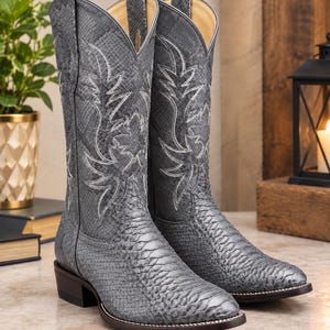May include: A pair of grey cowboy boots featuring a textured snakeskin pattern throughout the shaft and foot. The boots include white decorative stitching in a classic western design, pull tabs at the top, and a dark brown stacked heel.