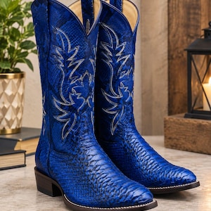 May include: A pair of vibrant blue cowboy boots featuring a textured snakeskin pattern throughout the shaft and foot. The boots include intricate silver-toned stitching in a leaf-like design on the upper shaft and a dark brown stacked heel.