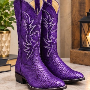 Puede incluir: Un par de botas vaqueras de color morado vibrante con un patrón de piel de serpiente texturizado, punta puntiaguda y tacón de bloque. La caña de cada bota está decorada con intrincadas costuras blancas en forma de hoja e incluye tiradores.