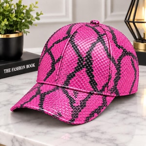Pode incluir: Um boné de beisebol com um padrão de pele de cobra em rosa vibrante e preto. O boné tem uma copa estruturada, uma aba curva e um pequeno botão rosa a condizer no topo central. O material apresenta uma textura semelhante a escamas.