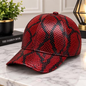 Pode incluir: Um boné de beisebol com um padrão texturizado vibrante em vermelho e preto, semelhante a pele de cobra. O boné tem uma copa estruturada, uma aba curva e um pequeno botão a condizer no centro do topo. O material parece brilhante e escamado.