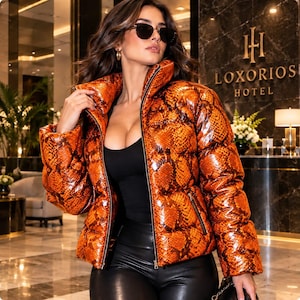 Peut inclure: Une doudoune brillante à motif peau de serpent orange et noir avec un col montant et une fermeture éclair intégrale. La veste présente des manches longues, une texture matelassée et deux poches latérales, portée sur un haut et un pantalon noirs.
