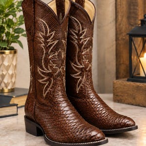 May include: A pair of brown cowboy boots featuring a textured snakeskin-like pattern across the exterior. The boots have a pointed toe, a low block heel, and intricate cream-coloured decorative stitching on the shaft in a leaf-like design.