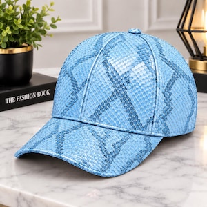 Pode incluir: Um boné de beisebol azul-claro com um padrão texturizado de pele de cobra em toda a superfície. O boné possui uma copa estruturada, uma aba curva e um pequeno botão a condizer no centro da parte superior.