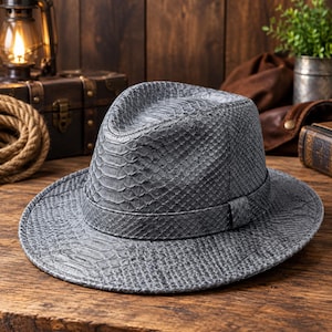 May include: A grey fedora hat featuring a textured snakeskin pattern across the entire surface, including the crown and the wide brim. The hat includes a matching grey snakeskin band wrapped around the base of the crown, creating a uniform look.