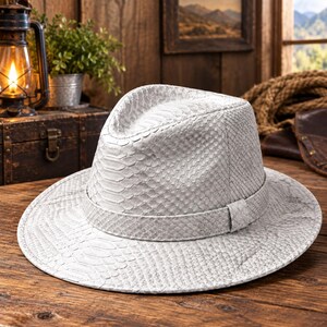 May include: A light gray fedora hat featuring a textured snakeskin pattern across the entire surface, including the crown and the wide brim. The hat includes a matching tonal band wrapped around the base of the crown, creating a uniform appearance.