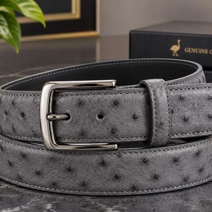Può includere: Una cintura grigia realizzata in pelle di struzzo testurizzata con un distinto motivo a pori su tutta la superficie. La cintura include una fibbia in metallo color argento lucido e un passante in pelle abbinato, dal design classico.