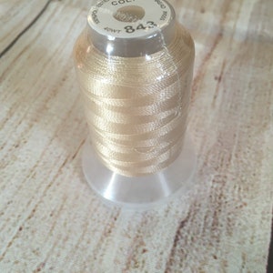 K&ouml;nnte beinhalten: Eine Spule hellbeige Stickgarn, mit der Nummer "843" und der Aufschrift "Polyester Embroidery Thread". Der Faden ist auf einer transparenten Kunststoffspule aufgewickelt. Der Hintergrund ist ein helles Holzmaserungsmuster.