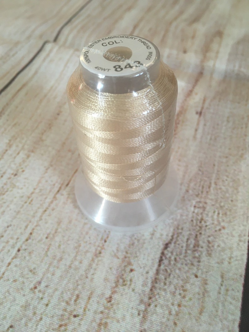 K&ouml;nnte beinhalten: Eine Spule hellbeige Stickgarn, mit der Nummer "843" und der Aufschrift "Polyester Embroidery Thread". Der Faden ist auf einer transparenten Kunststoffspule aufgewickelt. Der Hintergrund ist ein helles Holzmaserungsmuster.