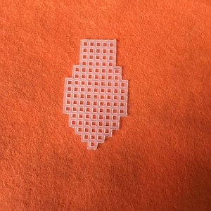 May include: A white plastic canvas shape, resembling a tie or decorative element, is centered on an orange felt background. The canvas is made of a grid of small, square holes. The background is a solid, vibrant orange.