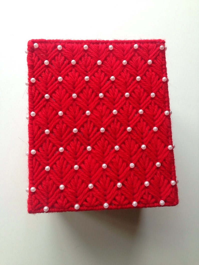 Op de afbeelding: Een vierkante, rode, met de hand gestikte doos met een textuurpatroon en witte parel kralen. De doos is gemaakt van garen en heeft een herhalend diamant- en bladerontwerp. De parels zijn gelijkmatig verdeeld.