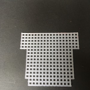 May include: A translucent, off-white plastic canvas sheet with a grid of square holes. The sheet is shaped like a stylized T-shirt, with a rectangular body and a shorter, wider top section. The edges are clean-cut.