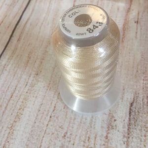 K&ouml;nnte beinhalten: Eine Spule hellbeige Ester-Stickgarn, Farbe 843, 40wt. Der Faden ist um eine transparente Kunststoffspule mit silbernem Deckel gewickelt. Der Hintergrund ist eine helle Holzmaserung.