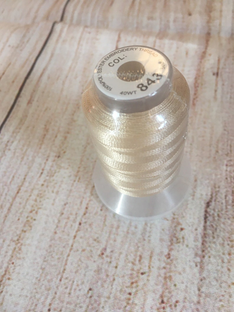 K&ouml;nnte beinhalten: Eine Spule hellbeige Ester-Stickgarn, Farbe 843, 40wt. Der Faden ist um eine transparente Kunststoffspule mit silbernem Deckel gewickelt. Der Hintergrund ist eine helle Holzmaserung.