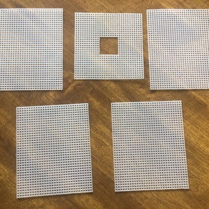 May include: Five white plastic canvas pieces in various rectangular shapes, including one with a square cut out of the center. The pieces are arranged on a wooden surface, showcasing a grid-like pattern.