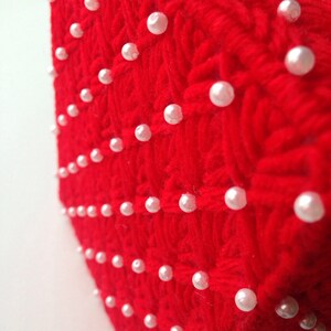 Op de afbeelding: Detailopname van een rood, getextureerd gebreid textiel, versierd met gelijkmatig verdeelde, witte parel kralen. Het textiel heeft een geweven patroon, en de parels zijn in nette rijen en kolommen gerangschikt.