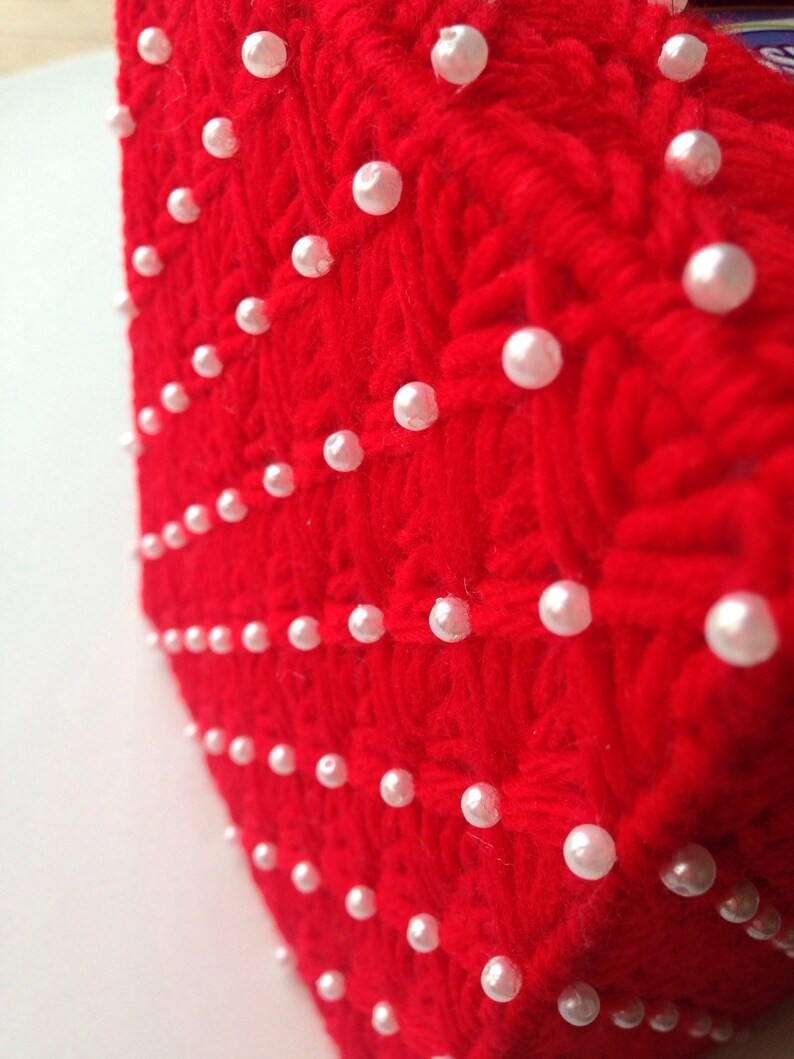 Op de afbeelding: Detailopname van een rood, getextureerd gebreid textiel, versierd met gelijkmatig verdeelde, witte parel kralen. Het textiel heeft een geweven patroon, en de parels zijn in nette rijen en kolommen gerangschikt.