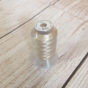 May include: A spool of light beige embroidery thread. The label reads "100% Polyester Embroidery Thread" and "COL: 843". The thread is wrapped around a clear plastic spool, sitting on a wooden surface.