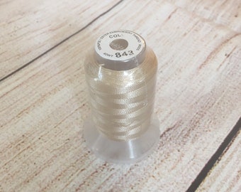 May include: A spool of light beige embroidery thread. The label reads "100% Polyester Embroidery Thread" and "COL: 843". The thread is wrapped around a clear plastic spool, sitting on a wooden surface.