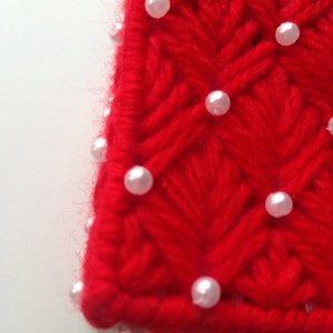 Op de afbeelding: Close-up van een rood, handgebreid textiel met een getextureerd, chevron-achtig patroon. Kleine, witte, parelachtige kralen zijn gelijkmatig verdeeld over het oppervlak. De randen van het textiel zijn enigszins vaag.