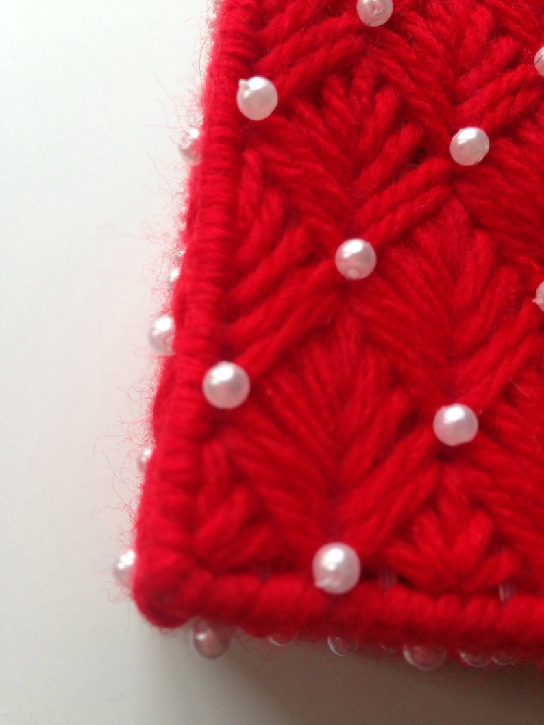 Op de afbeelding: Close-up van een rood, handgebreid textiel met een getextureerd, chevron-achtig patroon. Kleine, witte, parelachtige kralen zijn gelijkmatig verdeeld over het oppervlak. De randen van het textiel zijn enigszins vaag.