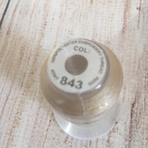 K&ouml;nnte beinhalten: Eine Spule beigefarbenes Stickgarn. Das Etikett lautet "100% Polyester Embroidery Thread" und "COL: 843". Die Spule steht auf einem transparenten Kunststoffsockel, der Hintergrund ist eine hellfarbige Holzoberfl&auml;che.