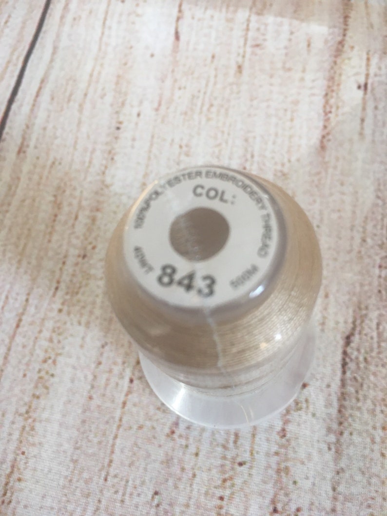 K&ouml;nnte beinhalten: Eine Spule beigefarbenes Stickgarn. Das Etikett lautet "100% Polyester Embroidery Thread" und "COL: 843". Die Spule steht auf einem transparenten Kunststoffsockel, der Hintergrund ist eine hellfarbige Holzoberfl&auml;che.