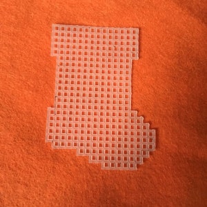 May include: A clear plastic canvas in the shape of a Christmas stocking, with a grid pattern. The stocking is placed on an orange felt background. This craft supply is used for needlepoint and cross-stitch projects.