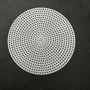 May include: A circular, off-white plastic canvas sheet with a grid pattern. The sheet is made of small, evenly spaced square holes arranged in concentric circles. The canvas is designed for crafting and needlework projects.