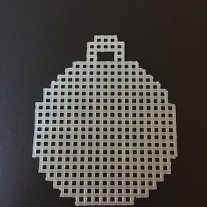 Op de afbeelding: Een helder, plastic canvas in de vorm van een kerstbal. Het canvas is gemaakt van een raster van kleine, vierkante gaten en heeft een kleine rechthoekige lus aan de bovenkant om op te hangen. De ornament is geplaatst tegen een zwarte achtergrond.